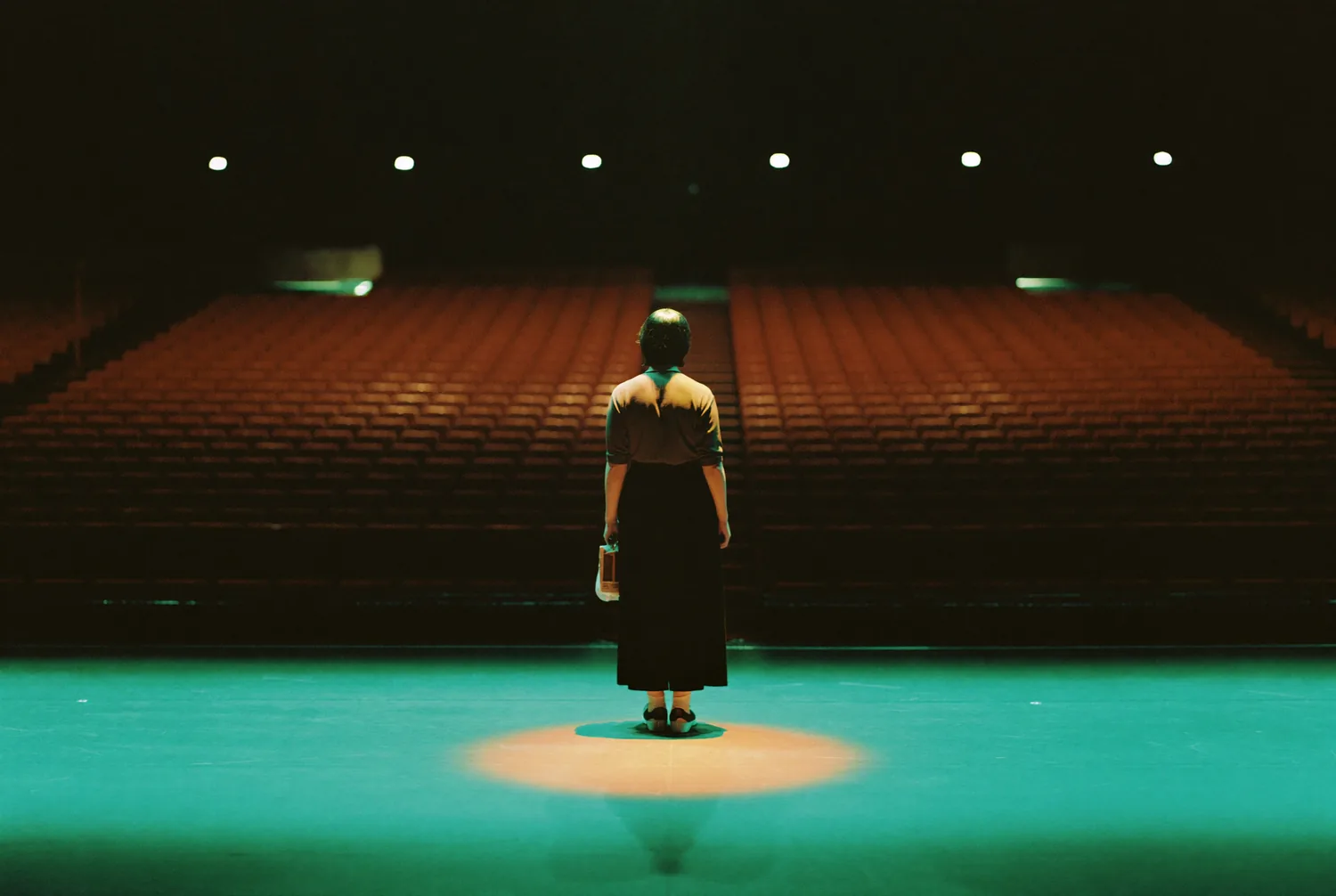A lone figure under a single spotlight on a stage with empty seats beyond A lone figure under a single spotlight on a stage with empty seats beyond