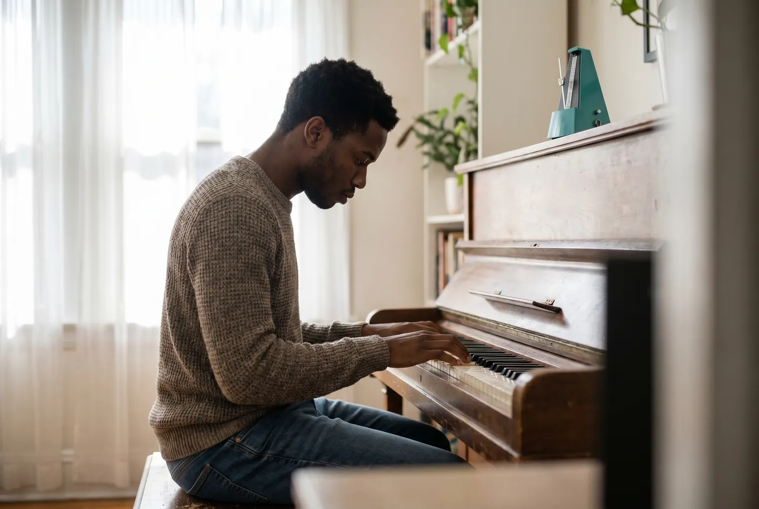 Young man practicing scales at an upright piano in morning light with a single teal metronome on the piano top Young man practicing scales at an upright piano in morning light with a single teal metronome on the piano top