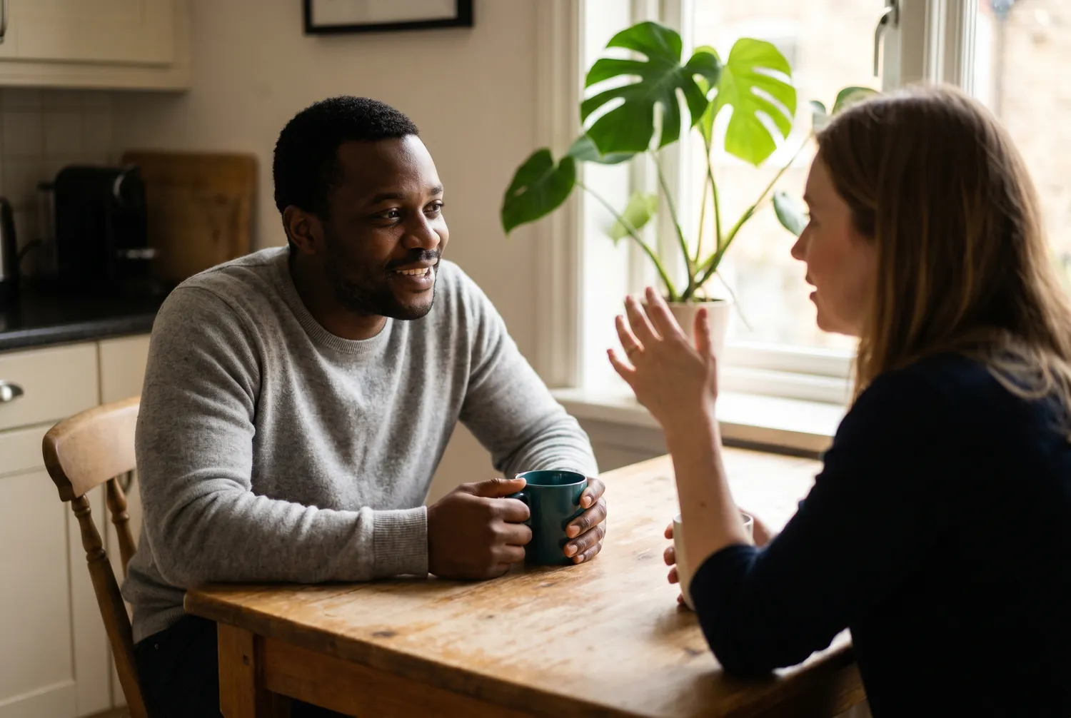 Two adults in genuine conversation at a sunlit kitchen table, one holding a deep teal mug Two adults in genuine conversation at a sunlit kitchen table, one holding a deep teal mug